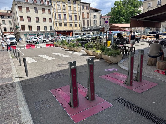 Square in France with some parked cars, but also a large terrace that is kept car free by the ways of blocking cars: plastic barriers, plant pots and metal spikes.