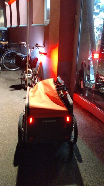 Ein rote-weisses Brompton-Faltrad mit einem angehängtem orange-graubraunem Croozer Cargo Lastenanhänger mit nicht ganz zugehendem Verdeck und einem darunter herausguckenden Fahrradsattel und Lenkstangengelenk eines gefaltetem Brompton-Faltrades steht vor einem erleuchteten Fahrradwerkstatt-Fenster.

Ansicht von hinten, Es sind zwei Rücklichter am Anhänger, eins an der Sattelstange des Zugfahrrades sowie ein Frontlicht (zum Gesehen werden) an der Lenksäule des Zugfahrrades sind an. Die letzten beiden beleuchten die Wand, an der es angelehnt ist.