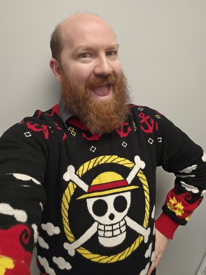 Selfie of a bald, thick bearded light skinned man wearing a black sweater that has the Straw Hat Crew from One Piece's Jolly Roger in the center (skull and crossbones wearing a straw hat). Assorted, tacky symbols are seen throughout in rows including red anchors, yellow devil fruit in red flames, and white clouds.