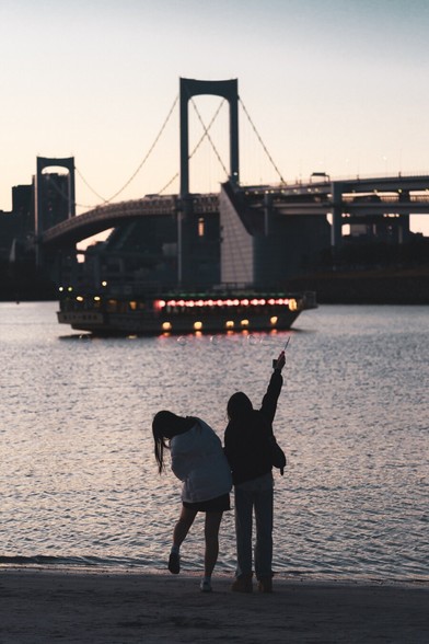 The image depicts two people near a body of water, engaging with bubbles, at dusk. In the background, a boat adorned with lights is visible, with a large bridge and a cityscape in silhouette. The scene has a tranquil and picturesque ambiance.