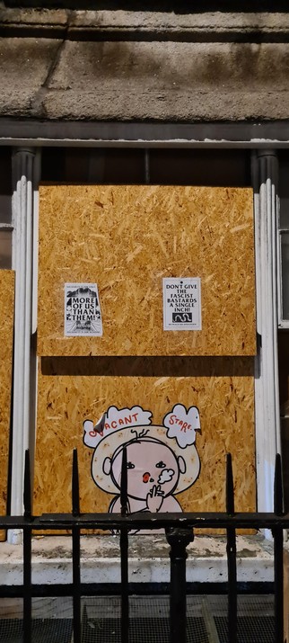 Boarded up, white, wooden framed window of the ground floor of a derelict vacant building behind a black iron bar fence with three posters:

Top left, from top to bottom, black letters on white, potrait oriented, A4-sized paper: Solidarity is our strenght, indistinct graphics surrounding the text more of us then them, solidarity is our weapon.

Top right, from top to bottom, black letters on white, potrait oriented, A4-sized paper: Don't give the fascist bastards an inch, an image of a white snake cut in pieces in a black square, No peace! No apologies!

Bottom middle: a large paper cut out of a sketch like, cartoonish, pink colored, round faced person wearing a cream coloured round hat. They are seen from mid torso upwards, touching the window sill in such a way it looks like they are looking out of the window. They are holding a cigaret, smoke escaping their red, puckered lips, while looking directly at the viewer with wide open eyes. Around the head two smoke clouds with the words vacant stare.