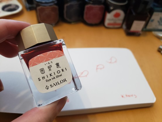 my hand, holding a bottle of orange Japanese ink. Behind it, is my sketchbook with today's drawing started on the open page.