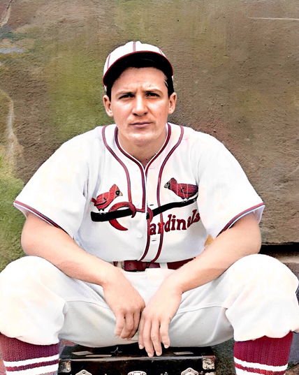 Colorized vintage photograph of baseball player Joe ‘Ducky’ Medwick in a St. Louis Cardinals uniform. He is sitting on a trunk against a muted wall background, arms resting on his knees. Medwick wears a classic white uniform with red trim, featuring two cardinal birds perched on a bat across the chest, and a matching cap. His expression is serious and focused.