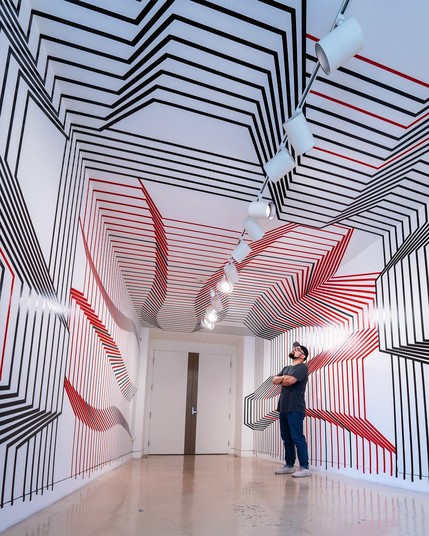 A white-walled gallery with high ceilings covered in black and red tape lines creating jagged and curving indentations across the walls and ceiling. A bearded man wearing a black tshirt and jeans is standing to the right of the room with arms crossed, looking upward.