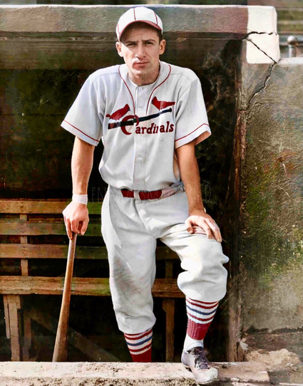 Colorized historical photograph of baseball player Joe “Ducky” Medwick in a St. Louis Cardinals uniform. He is standing in a dugout, holding a baseball bat in his right hand and resting his left hand on his bent left knee. He wears a white uniform with red trim, featuring two red cardinals perched on a black bat across the chest, and the word “Cardinals” in red script. He also wears a matching cap, red belt, and red-striped stirrup socks over white sanitary socks. The dugout walls are weathered and cracked, with wooden bench seating behind him.