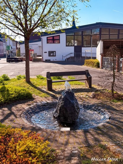 Ein idyllischer Dorfplatz in Laubach-Münster im Frühling: Im Zentrum plätschert ein kleiner Brunnen, umgeben von frischem Grün, einer schattigen Bank und traditionellem Naturstein. Sanftes Licht fällt durch das junge Laub der Bäume und verleiht dem Ort eine ruhige, friedliche Stimmung. Im Vordergrund ist das Dorfgemeinschaftshaus zu sehen, darüber ragt die Spitze des Kirchturms – stille Zeugen gelebten Dorflebens.