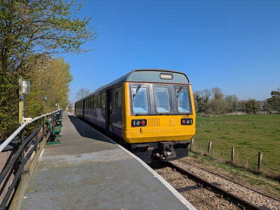 A two carriage BR Class 142 diesel multiple unit train stands at a country station platform in bright sunshine. The front is painted yellow, as all British trains should be.