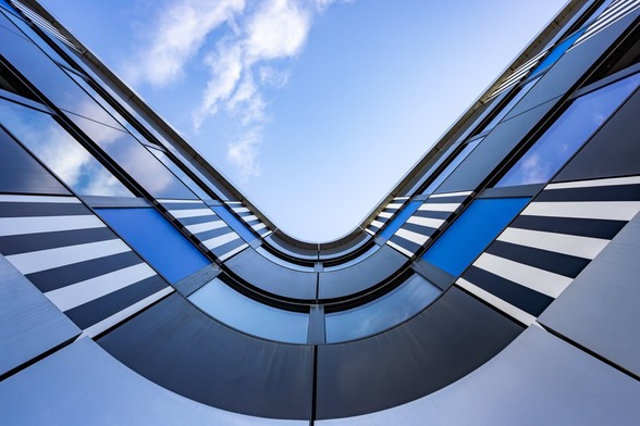 Look-up symmetrical view towards the sky from a curvy corner of a modern building decorated with panels in black, blue or white or with black and white stripes, in the Beaugrenelle quarter in Paris. The windows of the building reflect the blue skye and a few small white clouds.