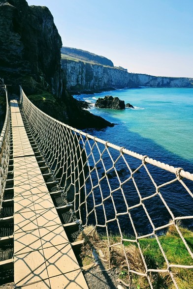 A long rope bridge stretches over turquoise water towards a rocky green island under a clear blue sky.