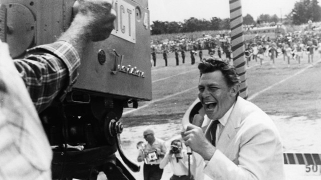 a  man in front of a camera wearing a white suit, talking excitedly into a microphone, a football field with a marching band on it behind him