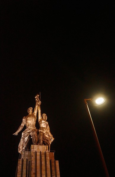 The illuminated Soviet monument “Worker and Kolkhoz Woman” stands out against the night sky. A lantern is placed next to it.