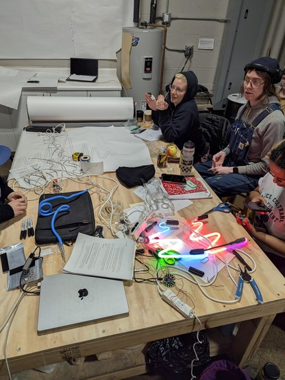 A very messy table with some students in the background. Two neon color samples, transformers, digitally drawn patterns, circuit playgrounds, notebooks and beverages covering the table