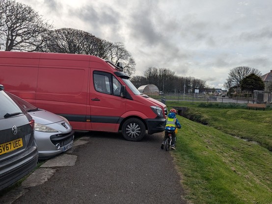 A child in hi-vis trying to cycle to nursery. A large red van is completely blocking the pavement.