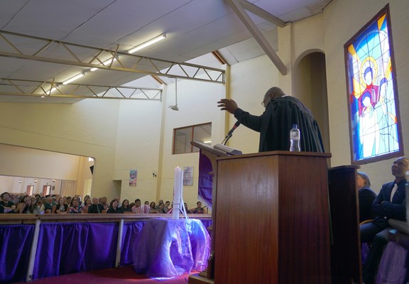 A preacher gestures from the pulpit, with a stained glass window behind to the right. The light from the window catches a tall candle on its purple clad table, in front of the rail. The congregation can be seen over the rail, under a high ceiling.