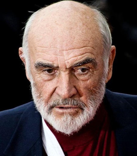 Bild von Sean Connery. Er trägt einen dunkelblauen Anzug und einen roten Rollkragenpullover. Der Hintergrund ist unscharf und zeigt dunkle Farben.