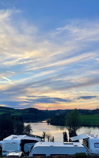 Ein malerischer Blick auf einen ruhigen See, umgeben von Grün und Hügeln, unter einem Himmel, der bei Sonnenuntergang mit weichen, bunten Wolken gefüllt ist. Zwei Wohnmobile sind im Vordergrund geparkt. 
A scenic view featuring a calm lake surrounded by greenery and hills, under a sky filled with soft, colorful clouds at sunset. Two campervans are parked in the foreground.