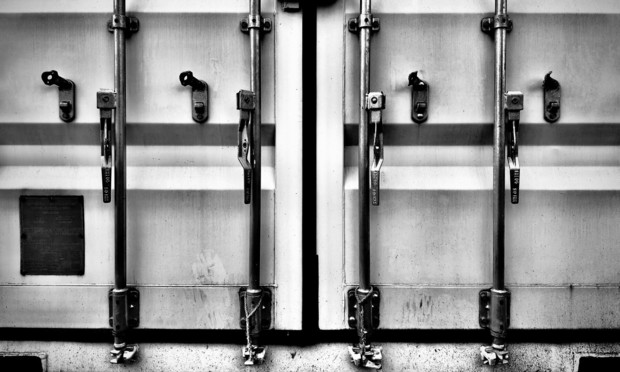 The large doors and vertical bar locks of a metallic container stand closed in slightly dirty shades of gray.