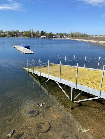 A tranquil reservoir scene featuring a yellow dock extending into the water. Two ducks swim near the shore, with visible submerged tires in the clear water. The background showcases green trees and buildings under a bright blue sky. On the right the dam wall is clearly visible well above the current waterline.