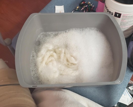 Gray basin with water, suds and white lace scarf in it, sitting on a chair with clutter