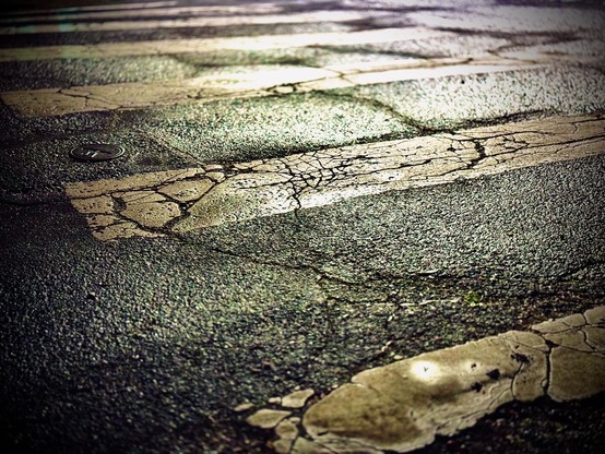 A city crosswalk with cracks in the white paint and bare road reflects a street lamp on its wet surface at night.