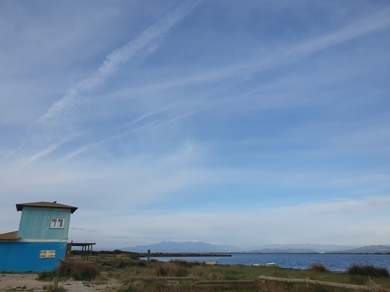 a blue wooden building next to a stretch of water. beyond a line of mountains, one, the Canigú still covered with snow. sky is blue with shapeless clouds