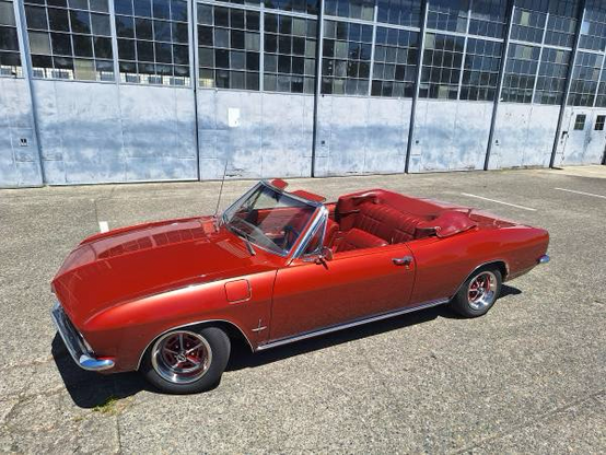 A 1966 Chevrolet Corvair Monza convertible with the top down, showing off its matching Aztec Bronze (orangey copper) paint and upholstery in a sunny parking lot.