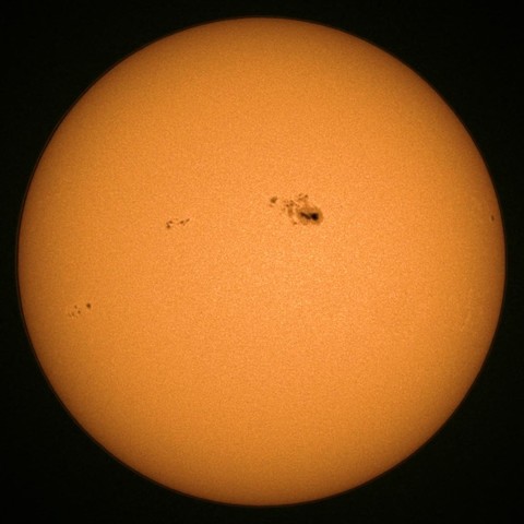 A close-up image of the Sun, showing its bright orange surface with several sunspots visible as dark patches.