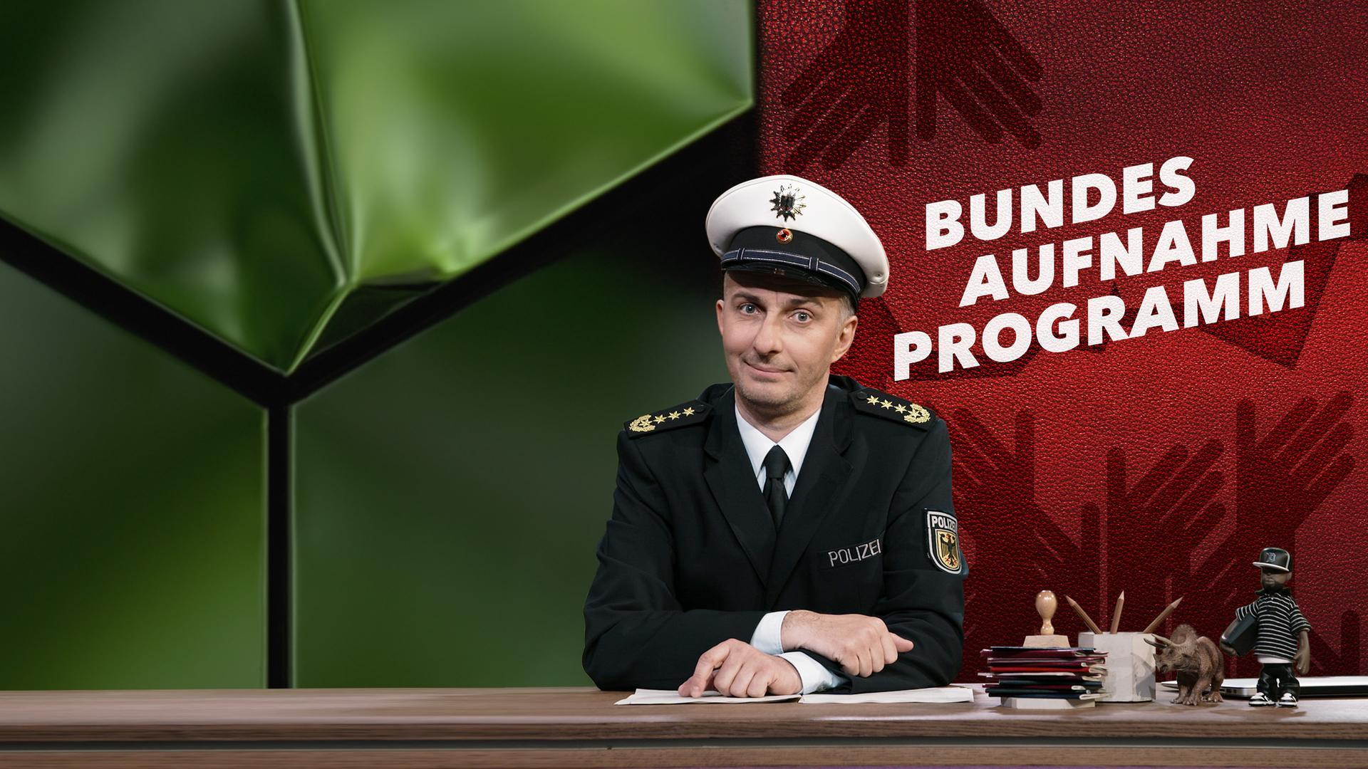 Jan Böhmermann sitzt in einer Polizeiuniform am Schreibtisch. Rechts neben ihm ist das Wort "Bundesaufnahmeprogramm" zu sehen.